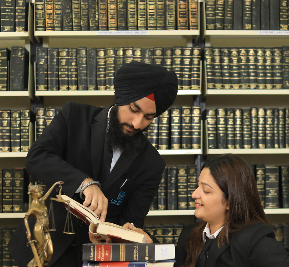 Two students in a library, one pointing at a book, surrounded by bookshelves.